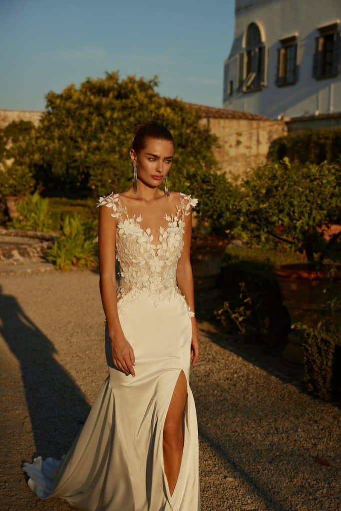 Trouwjurk met stretch satijnen rok, 3D bloemen, illusie halslijn, capmouwen en open achterkant met tulle strik voor een moderne en elegante uitstraling.