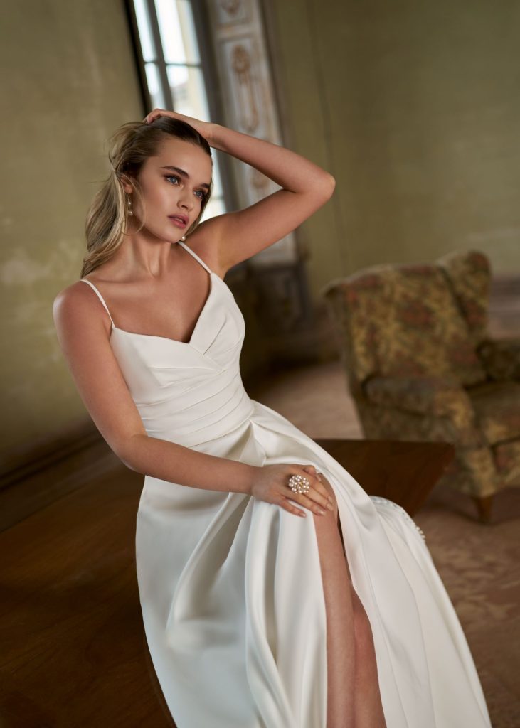 Satijnen A-lijn trouwjurk met geplooid lijfje, dunne bandjes, hoge split en minimalistisch modern design.